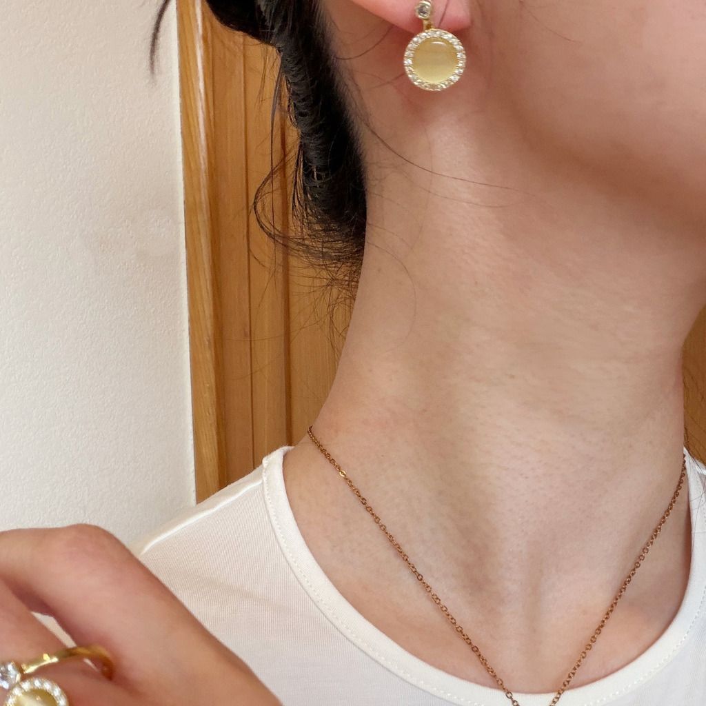 Close-up of a person wearing a round gold drop earring with a textured bezel and a delicate gold chain necklace, with a gold ring also visible.