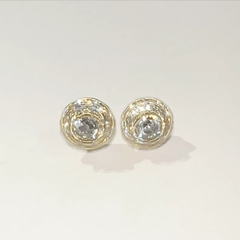 A pair of gold-toned stud earrings with a textured, woven-like circular design surrounding a clear faceted center stone, shown on a plain light background.