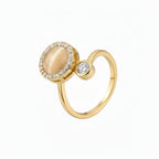A gold ring featuring a large oval cream cabochon surrounded by a halo of small clear stones, with a smaller round clear stone on the side, set against a plain white background.