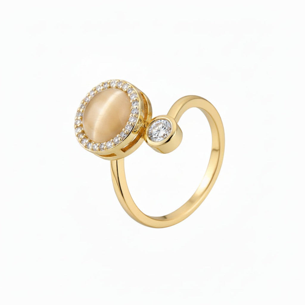 A gold ring featuring a large oval cream cabochon surrounded by a halo of small clear stones, with a smaller round clear stone on the side, set against a plain white background.