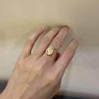 A gold ring with a round yellow gemstone and a small clear accent stone, worn on a person's hand against a plain beige background.