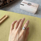 A silver ring with a round clear gemstone is worn on a person's finger, resting on a textured yellow-green mat. A white product box labeled 'Sakura' is visible in the background on a gray surface.