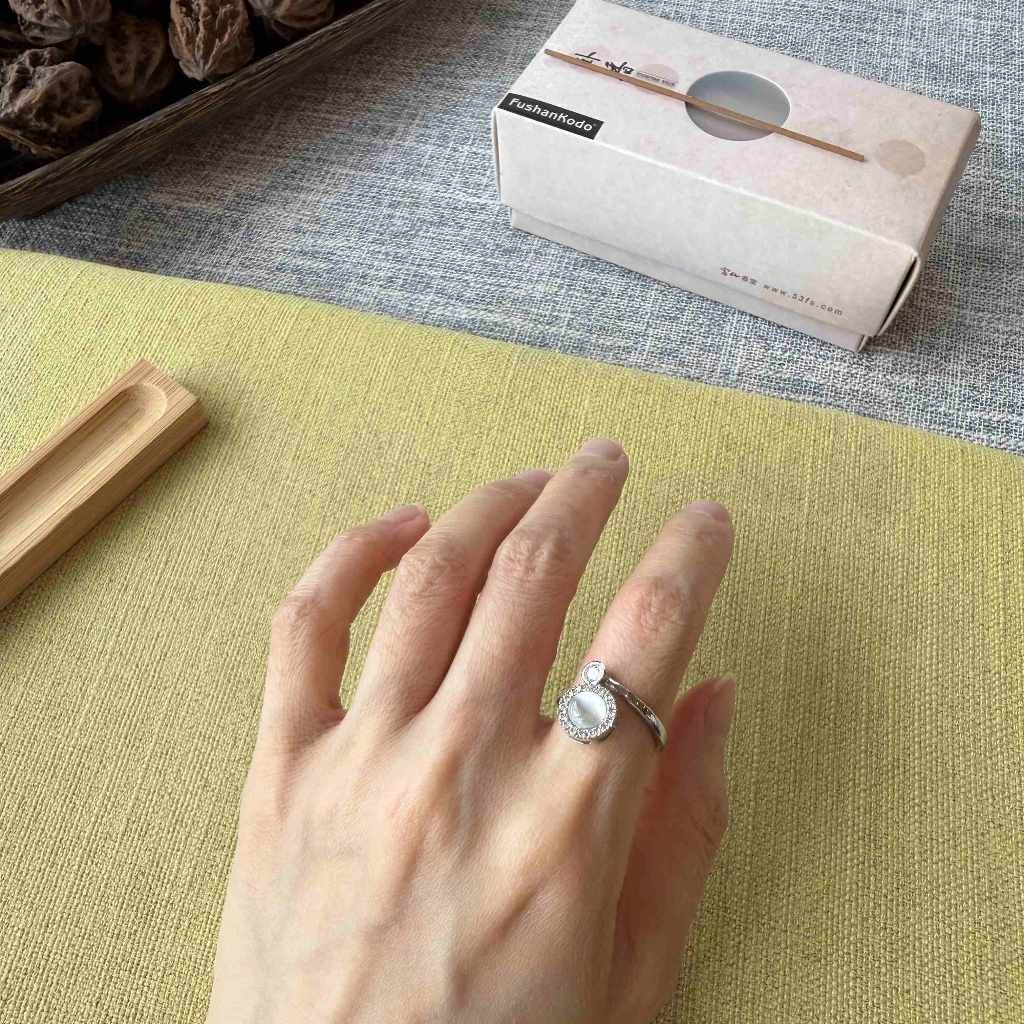 A silver ring with a round clear gemstone is worn on a person's finger, resting on a textured yellow-green mat. A white product box labeled 'Sakura' is visible in the background on a gray surface.
