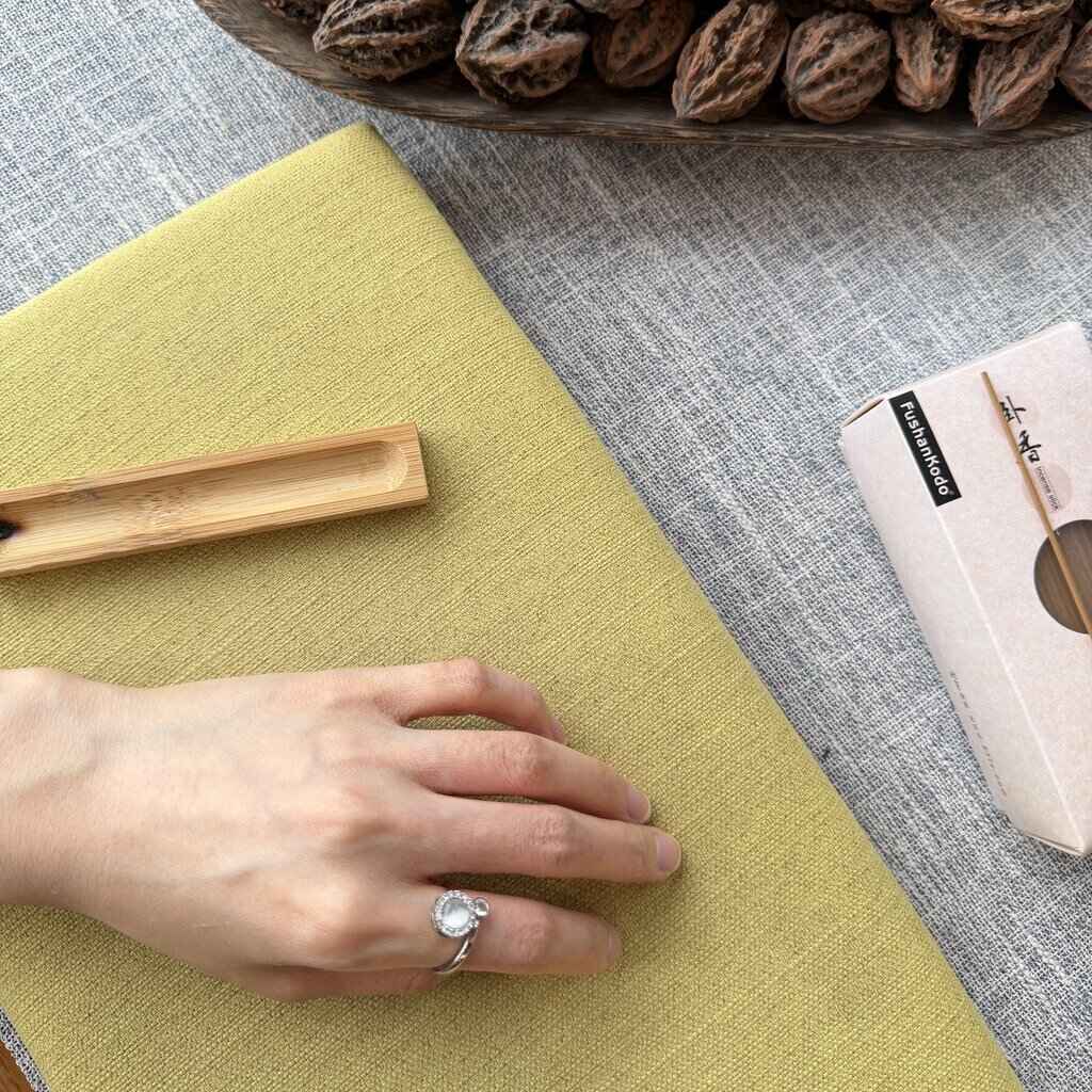 A close-up of a hand resting on a textured sage green fabric surface, with a bowl of walnuts and a small box in the background on a gray woven tablecloth.