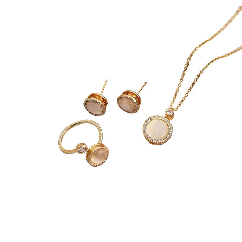 Gold jewelry set featuring a necklace with a round cream-colored stone surrounded by clear crystals, matching stud earrings, and a ring with a curved band and a small clear stone, all on a plain white background.