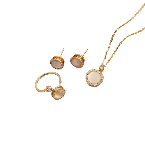 Gold jewelry set featuring a necklace with a round cream-colored stone surrounded by clear crystals, matching stud earrings, and a ring with a curved band and a small clear stone, all on a plain white background.