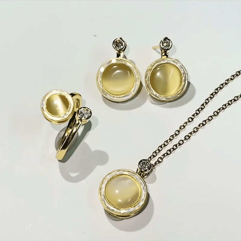A jewelry set featuring a gold pendant necklace, matching stud earrings, and a ring, all with round cream-colored cabochon stones and small clear crystal accents, displayed on a white background.