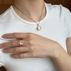 A close-up of a person wearing a delicate gold necklace with a round cream-colored pendant and a matching gold ring on their finger, both displayed against a white t-shirt.