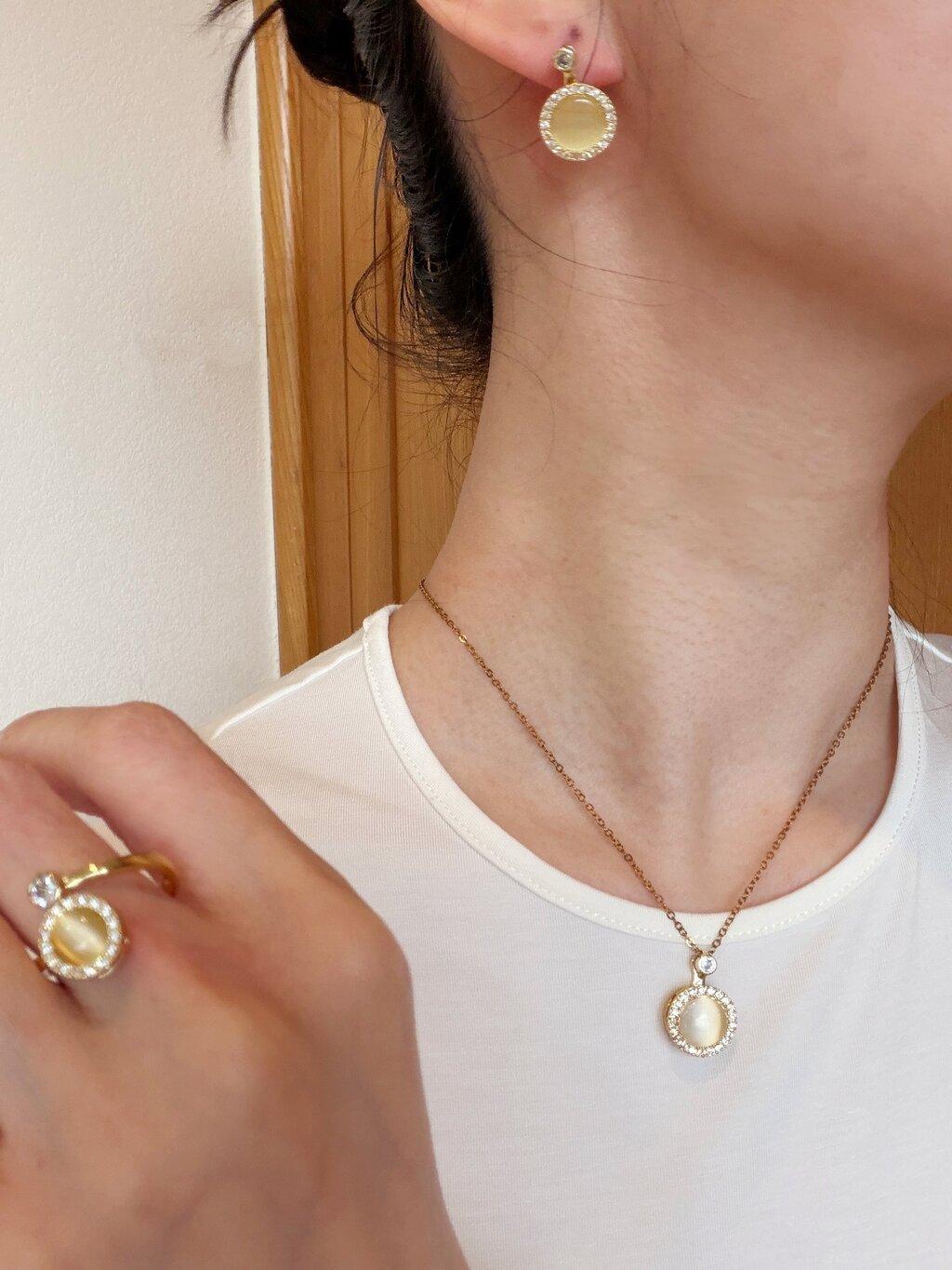 A close-up lifestyle shot of a model wearing a gold necklace with a round pendant and matching gold hoop earrings, along with a gold ring.