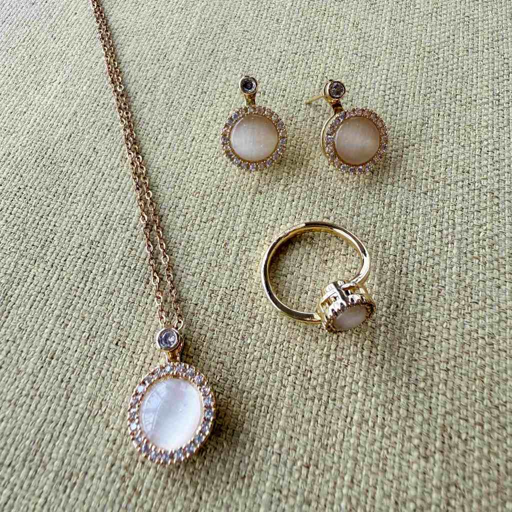 A gold jewelry set featuring a pendant necklace, matching stud earrings, and a ring, all with oval milky white stones surrounded by small clear crystals, displayed on a textured beige fabric background.