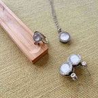 Silver-toned jewelry set featuring a ring, pendant necklace, and stud earrings with round white stone centers, displayed on a textured beige fabric surface with a wooden block prop.