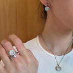 A close-up lifestyle shot of a person wearing a silver-toned ring with a round iridescent shell centerpiece and a matching silver necklace with a round pendant, against a light wood background.