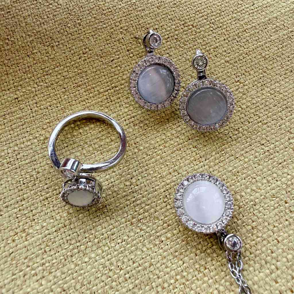 A jewelry set featuring a silver-toned ring and matching earrings with oval milky white stones surrounded by small clear crystals, displayed on a textured beige fabric background.