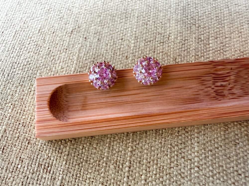 A pair of round stud earrings covered in small pink rhinestones, displayed on a wooden stand over a beige woven fabric background.
