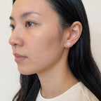 A close-up side profile of a woman wearing a small, round, clear gemstone stud earring on her earlobe against a plain light beige background.