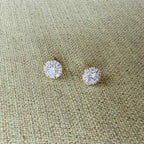 A pair of round stud earrings with clear gemstones in a gold-tone halo setting, displayed on a textured beige woven fabric background.