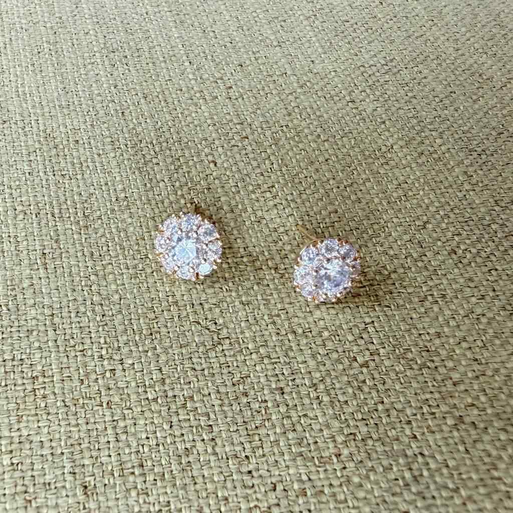 A pair of round stud earrings with clear gemstones in a gold-tone halo setting, displayed on a textured beige woven fabric background.