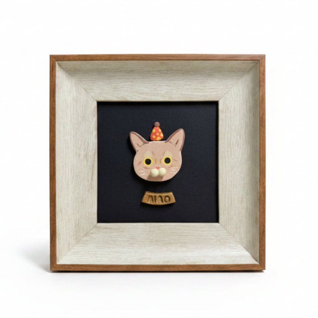 A square framed miniature wooden cat figurine wearing a small orange hat and a gold collar with the text 'AM10'. The cat is mounted on a black backing inside a light-colored wood frame, set against a plain white background.
