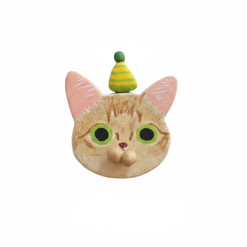A novelty cat head mask or face covering featuring an orange tabby cat design with pink ears, large green eyes, and a small green and yellow striped party hat on top, shown against a plain white background.