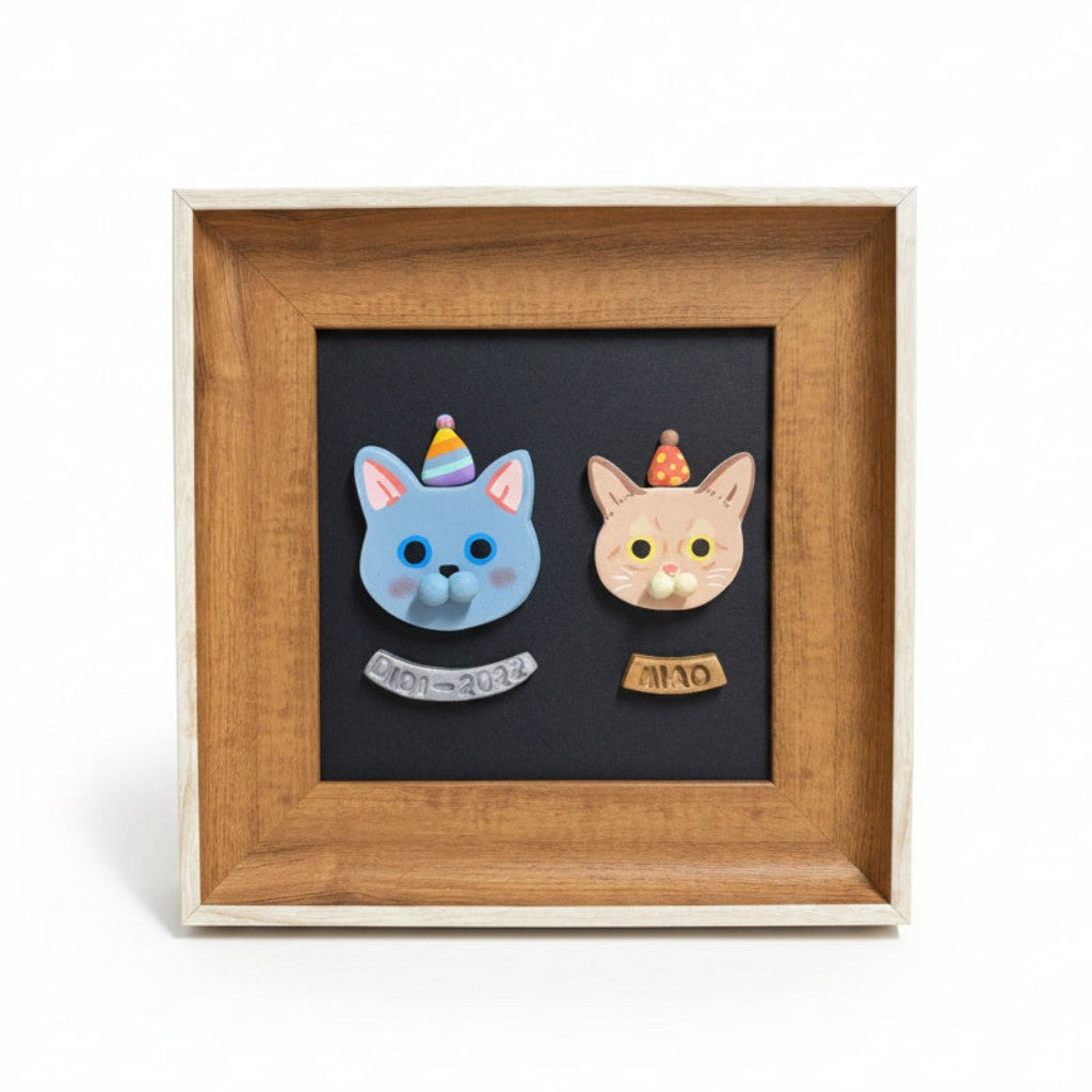 A square wooden picture frame containing a black background with two small cat head cutouts wearing party hats and small labels, photographed on a clean white background.