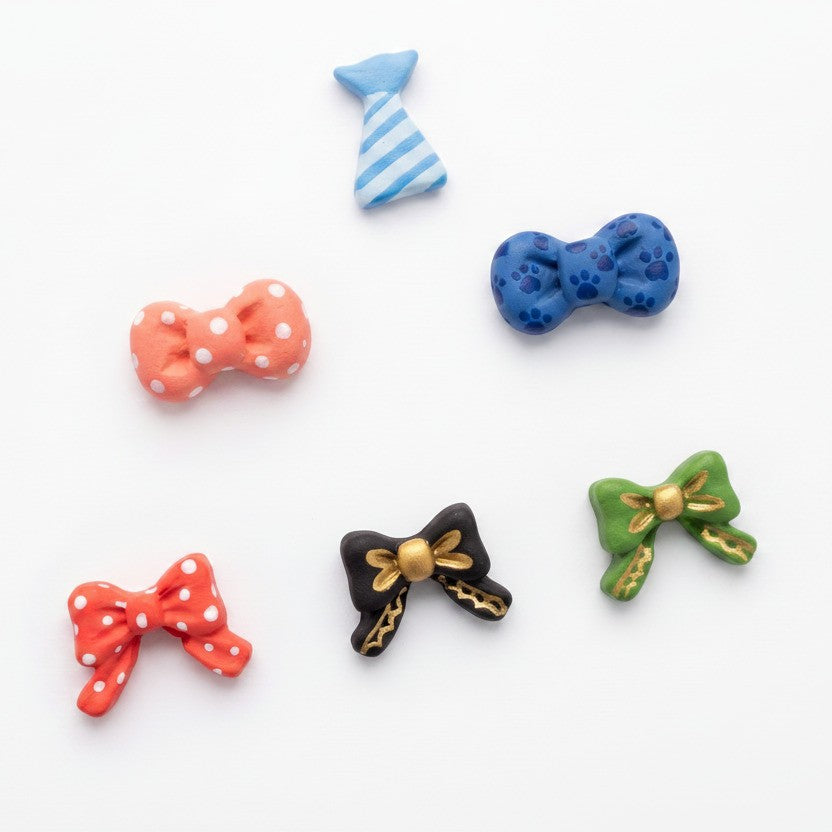 Six small bow-shaped accessories in assorted colors and patterns are arranged on a plain white background. The set includes a blue-and-white striped bow, a blue paw-print patterned bow, an orange polka-dot bow, a red polka-dot bow, a black bow with gold trim, and a green bow with gold trim.