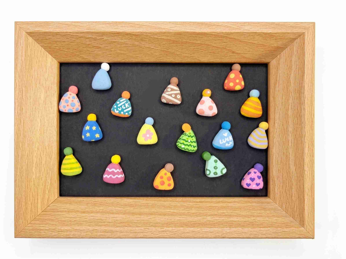 A square wooden frame containing a black background with multiple small, colorful, patterned hat-shaped items arranged on it, photographed against a plain white background.