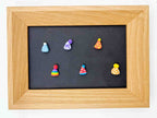 A square wooden frame with a black center panel displaying six small colorful party hat charms or magnets arranged in two rows, photographed against a plain white background.
