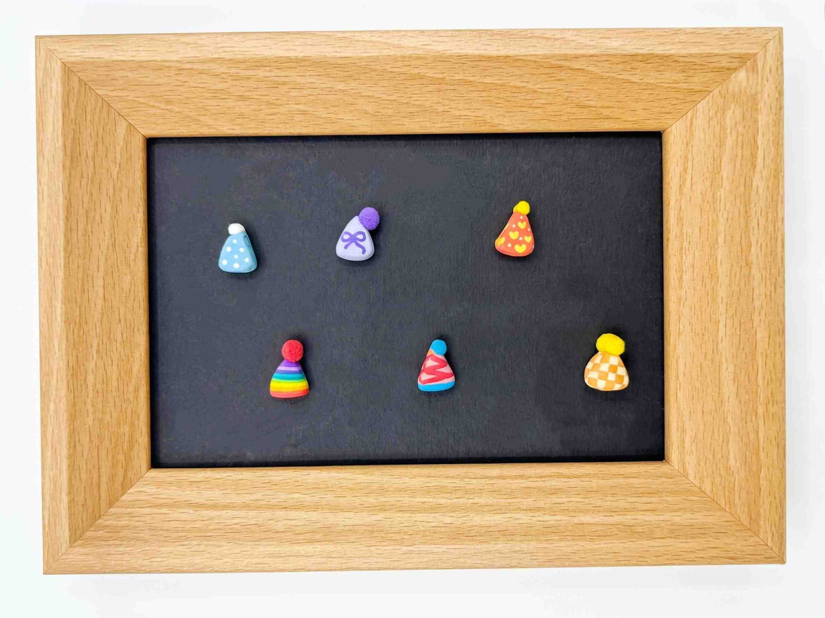 A square wooden frame with a black center panel displaying six small colorful party hat charms or magnets arranged in two rows, photographed against a plain white background.