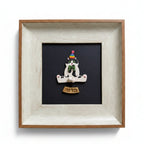 A square framed art piece featuring a small white cat figurine sitting cross-legged on a black background, wearing a colorful party hat and green bow tie, with a small wooden plaque below. The piece is housed in a light natural wood frame against a white background.