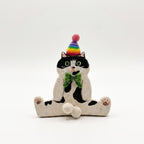 A ceramic figurine of a black and white cat sitting cross-legged, wearing a rainbow-striped party hat and a green bow tie, on a seamless white background.