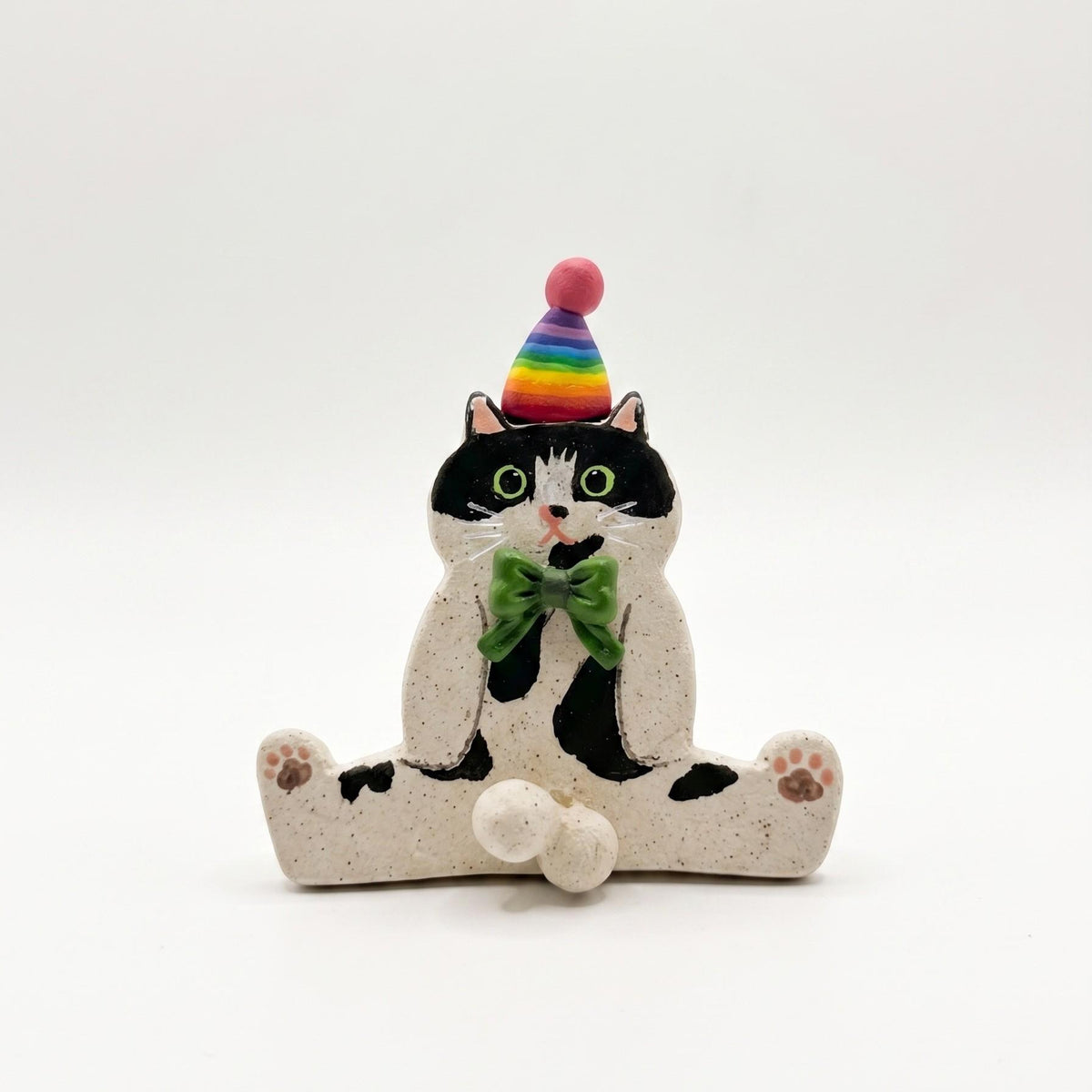 A ceramic figurine of a black and white cat sitting cross-legged, wearing a rainbow-striped party hat and a green bow tie, on a seamless white background.