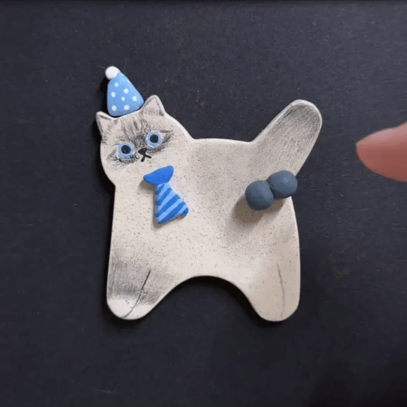 A handmade ceramic cat-shaped plaque or figurine on a dark background, featuring a gray cat body, blue party hat, blue striped tie, and blue paw pads. A human finger is visible on the right edge.