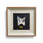 A framed art piece featuring a small white ceramic dog figurine wearing a blue hat and scarf, mounted on a black background with a yellow 'OTOP' label. The piece is housed in a square frame with a light-colored wood and white textured finish, set against a plain white background.