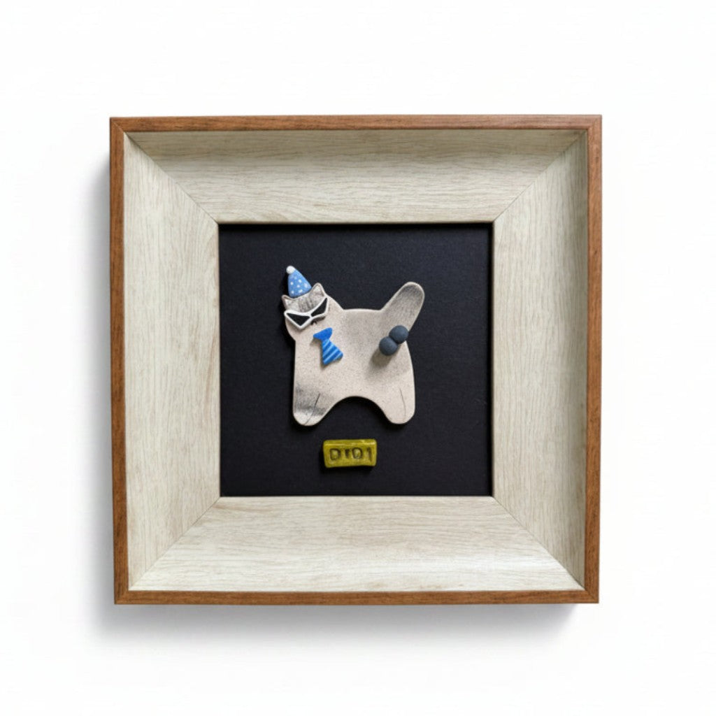A framed art piece featuring a small white ceramic dog figurine wearing a blue hat and scarf, mounted on a black background with a yellow 'OTOP' label. The piece is housed in a square frame with a light-colored wood and white textured finish, set against a plain white background.