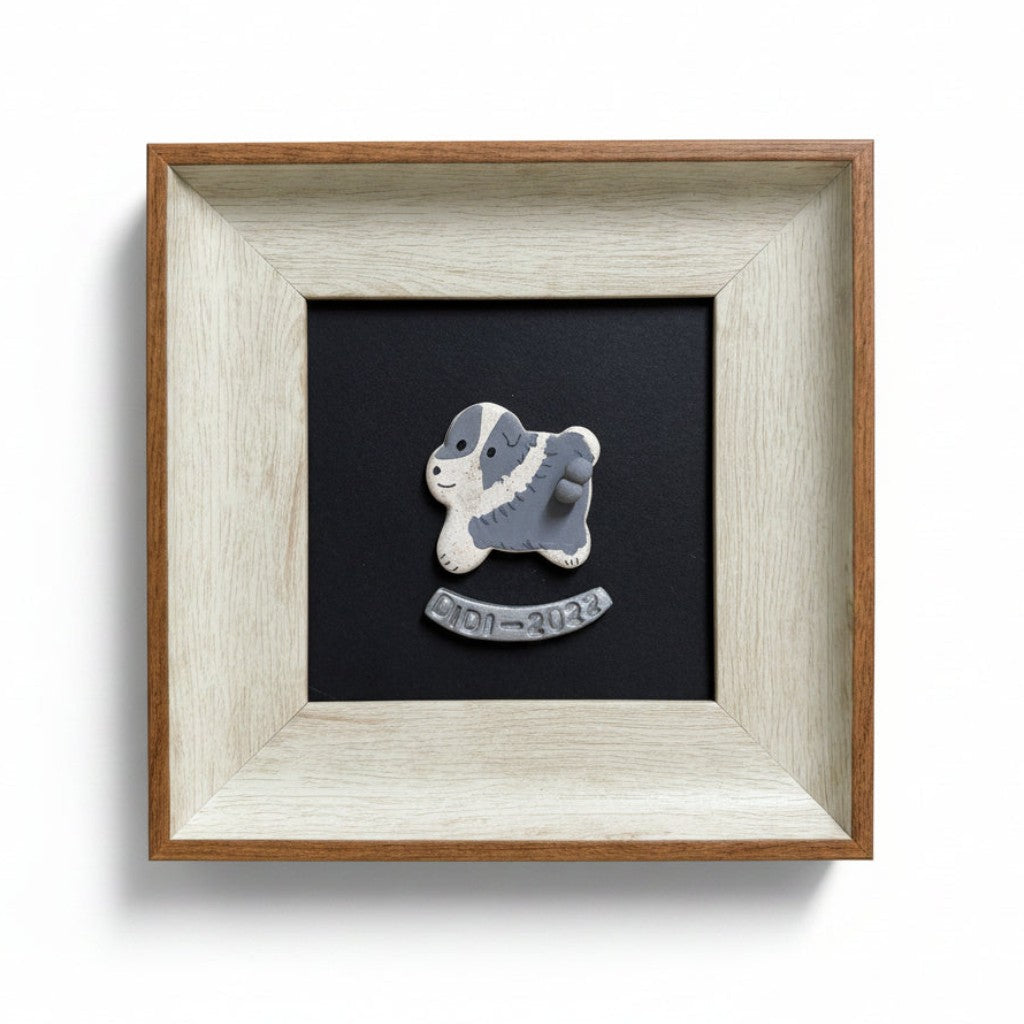 A square framed memorial plaque featuring a small gray and white dog figurine on a black background, set in a light natural wood frame against a white wall.