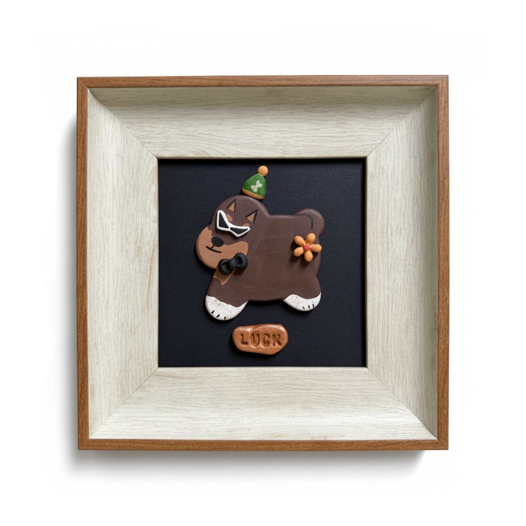 A square framed decorative art piece featuring a brown cartoon dog ornament with white paws, a green party hat, and an orange flower, mounted on a black background inside a light wood and off-white frame. A small brown tag below the dog reads 'LOVE'.