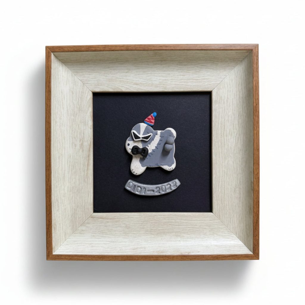 A custom wooden shadow box frame featuring a gray and white dog figurine wearing sunglasses and a party hat, mounted on a black background with the name 'DIDI - 2020' engraved on a banner. The frame is set on a light wood desk with a color chart in the background.