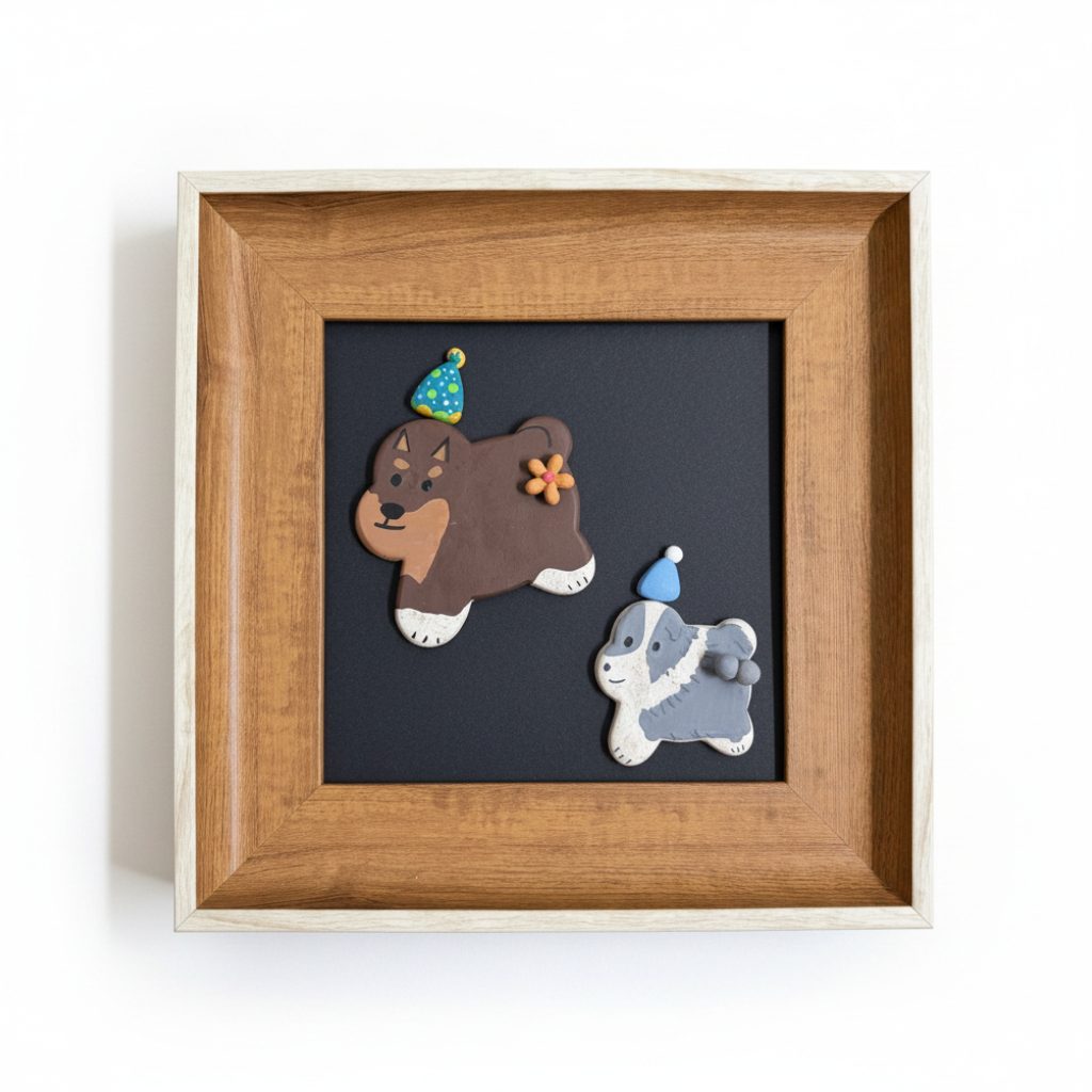 A square wooden shadow box frame containing a dark background with two small dog figurines wearing party hats and a small orange flower accent, photographed against a plain white wall.