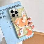 A hand holds a clear phone case decorated with cute cartoon cat illustrations and a 3D orange cat pop socket. The phone case is shown on a light blue phone, resting on a light blue book with handwritten text.