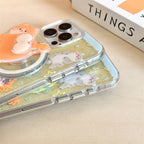 A clear phone case with a pastel cat illustration and a built-in pop socket, shown on a light wood surface next to a white book.