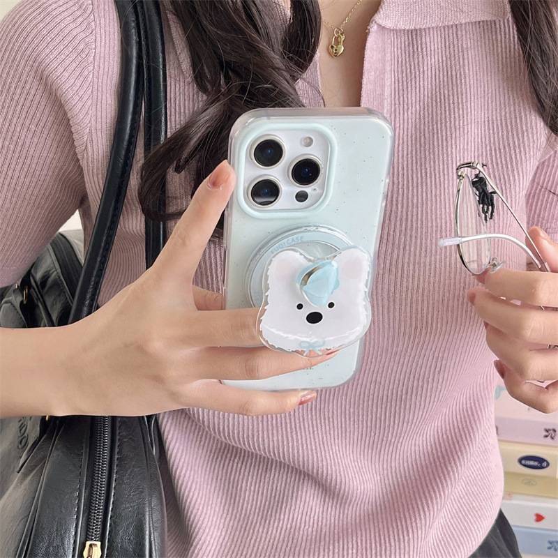 A close-up lifestyle shot of a person holding a light blue phone case with a white bear-shaped pop socket. The person is wearing a pink ribbed knit top and holding a black shoulder bag and eyeglasses.