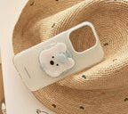 A clear phone case with a cute 3D bear face accessory, displayed on a white surface next to a woven straw hat and eyeglasses.