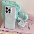 A clear phone case with light blue speckles and a circular MagSafe ring, shown on a white phone. The phone is styled on pink books next to a mint green padded cup and a small bear-shaped accessory.
