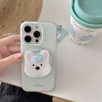 A hand holds a light gray speckled phone case featuring a cute 3D bear face design with pink ears, a blue bow, and a clear strap. A matching cup and woven hat sit on a white table in the background.