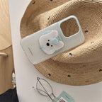 A clear phone case with a cute 3D bear face design, displayed on a woven straw hat with eyeglasses nearby.