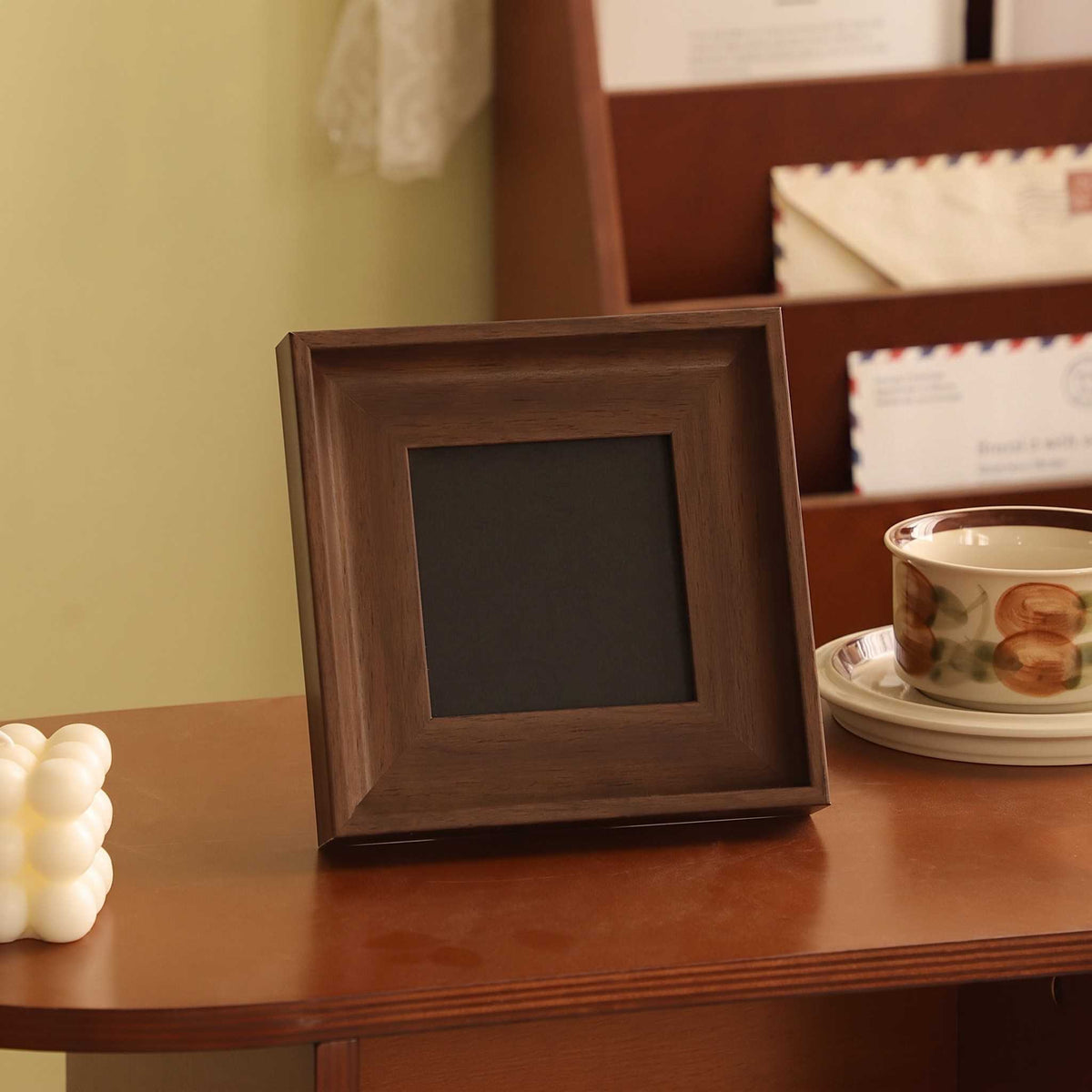 A square dark brown wooden picture frame with a black center, standing on a wooden table next to a ceramic cup and saucer.