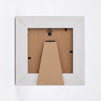 The back of a square picture frame in white, showing the brown cardboard backing and mounting hardware against a white background.