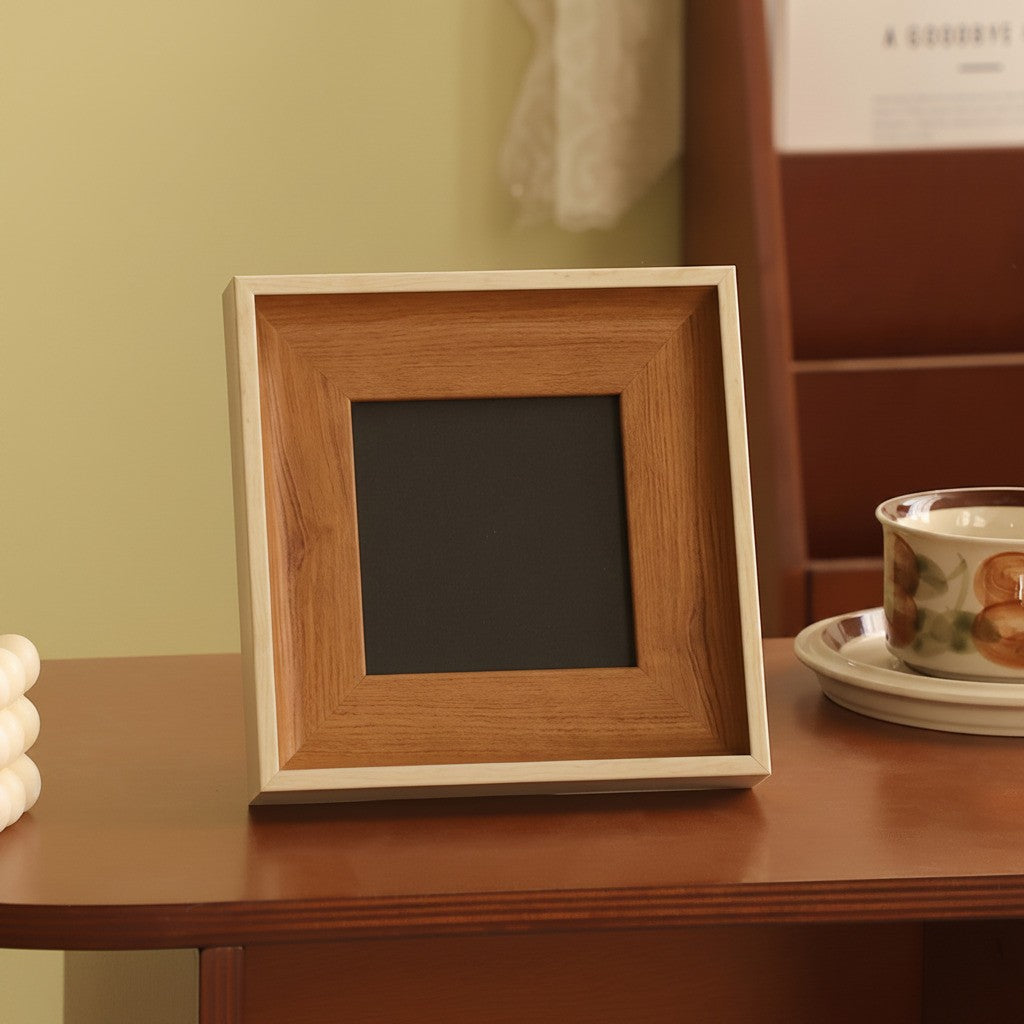A square picture frame with a warm medium-brown wood grain outer border and a light beige inner frame, standing on a wooden table next to a teacup.