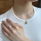 A close-up lifestyle shot of a person wearing a delicate rose gold chain necklace with a flower-shaped pendant and a matching rose gold ring with a clear crystal cluster on the finger, set against a white t-shirt and a wooden background.