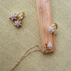 A gold jewelry organizer made of a bamboo wood block with three burnished holes, displayed on a beige linen background. The organizer is shown holding a gold ring, a gold necklace, and a pair of gold snowflake stud earrings.
